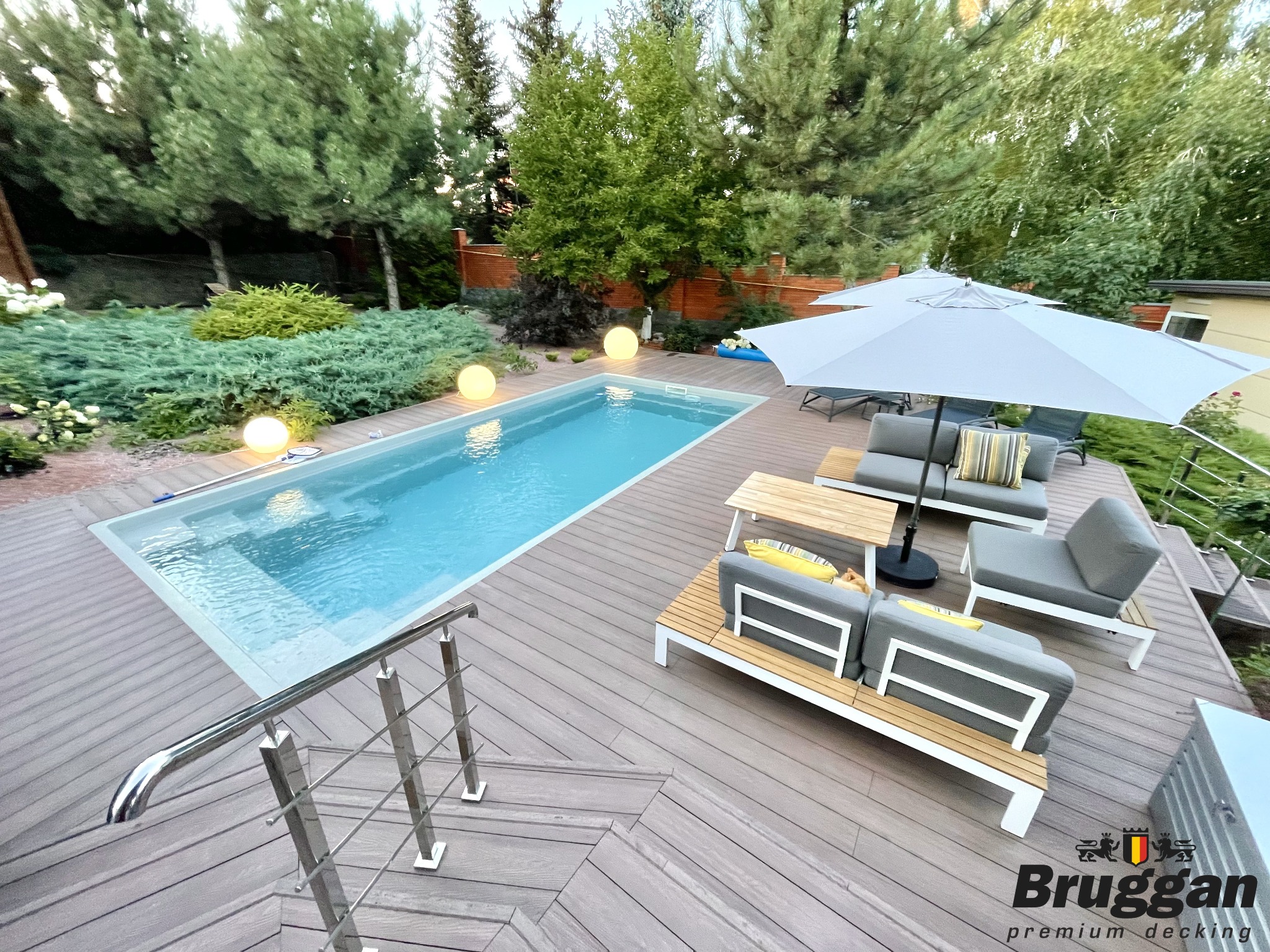 Nowoczesny taras z desek kompozytowych w odcieniach szarości, przylegający do basenu z oświetleniem kulistym, z meblami ogrodowymi i schodami z metalową barierką.