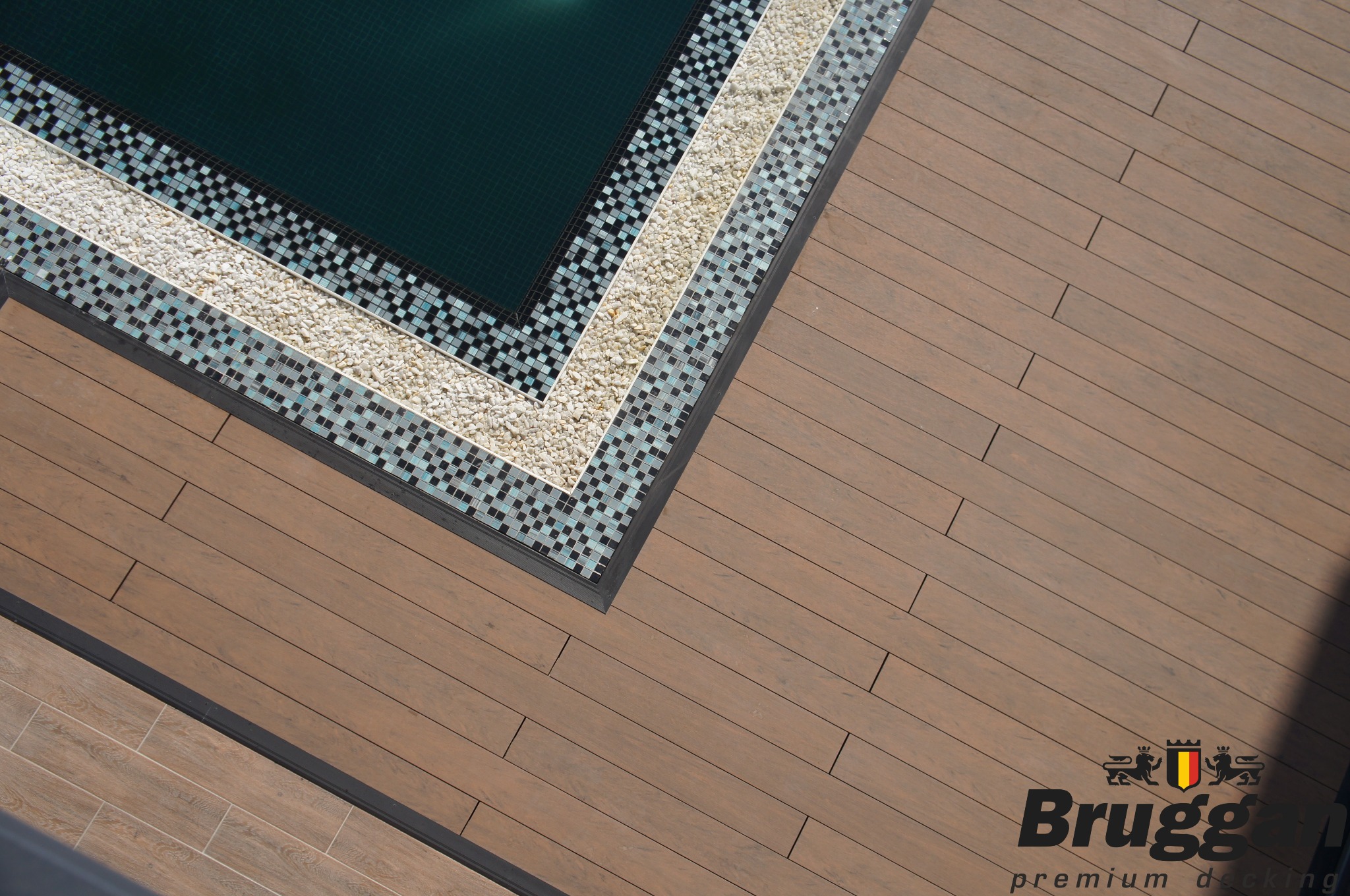 Detale wykończenia tarasu z desek kompozytowych przy basenie z mozaiką i białymi kamykami dekoracyjnymi, widok z góry, logo producenta desek w rogu.