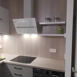 Szara kuchnia z drewnianym blatem i płytą indukcyjną, biały okap i półki z pojemnikami. Pralka zabudowana w szafce. Oświetlenie podszafkowe.