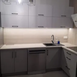 Jasnoszara kuchnia z drewnianym panelem ściennym i czarnymi uchwytami. Nad szafkami wiszą trzy lampy w czarnych, geometrycznych oprawach.