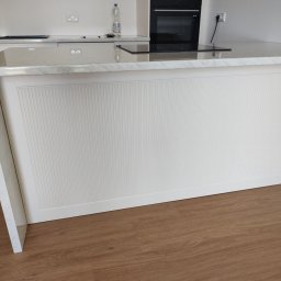 AWANGARDE - Biała wyspa kuchenna z frezowanymi frontami na tle zabudowy kuchennej z piekarnikiem Bosch i marmurowym blatem, na podłodze panele drewnopodobne.