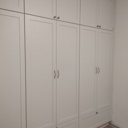 AWANGARDE - Biała, zabudowana szafa z szufladami i górnymi szafkami, z delikatnymi uchwytami, na tle jasnej ściany i drewnianej podłogi ułożonej w jodełkę.