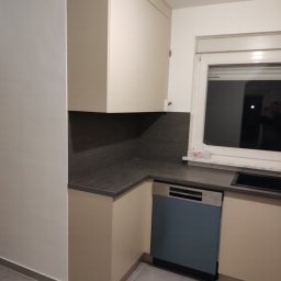 AWANGARDE - Jasna kuchnia z szarym blatem i niebieską zmywarką w zabudowie. Ujęcie z niskiego kąta, widoczny fragment okna i szare płytki podłogowe.