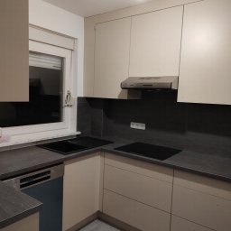 AWANGARDE - Jasna kuchnia z szarym blatem i niebieską zmywarką. Widoczna płyta indukcyjna, okap i gładkie fronty szafek. Minimalistyczny design, stonowana kolorystyka.