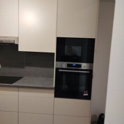 AWANGARDE - Jasna zabudowa kuchenna z piekarnikiem i płytą w Koninie. Minimalistyczne fronty szafek, szary blat i płytki nad blatem. Widoczna refleksja w szybie piekarnika.