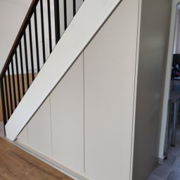 AWANGARDE - Biała szafa wnękowa pod schodami z czarną balustradą. Drewniana podłoga i szare płytki tworzą nowoczesny kontrast w jasnym wnętrzu.