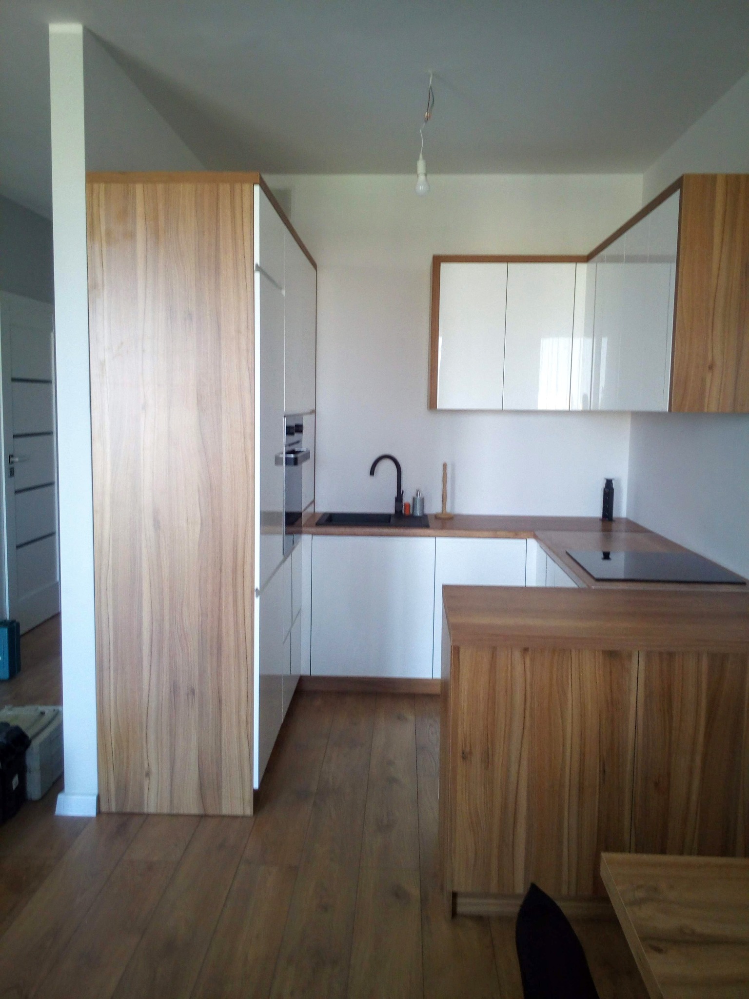 Nowoczesna kuchnia z białymi szafkami i drewnianymi frontami, czarny zlew i płyta indukcyjna, drewniana podłoga i stolik w tle, surowa żarówka zwisająca z sufitu.