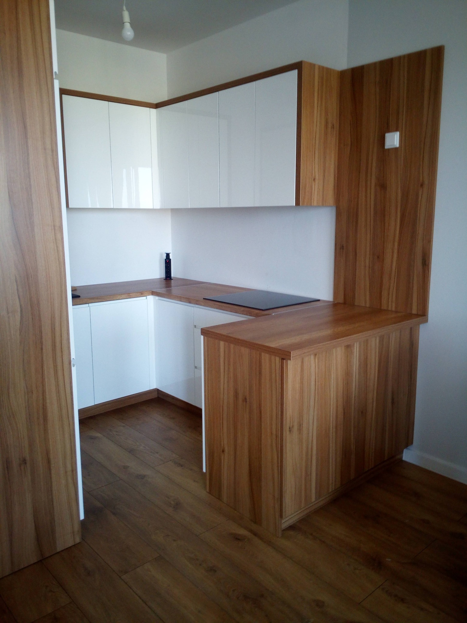 Nowoczesna kuchnia z białymi szafkami górnymi i dolnymi, drewnianym blatem i panelem bocznym z widoczną płytą indukcyjną.