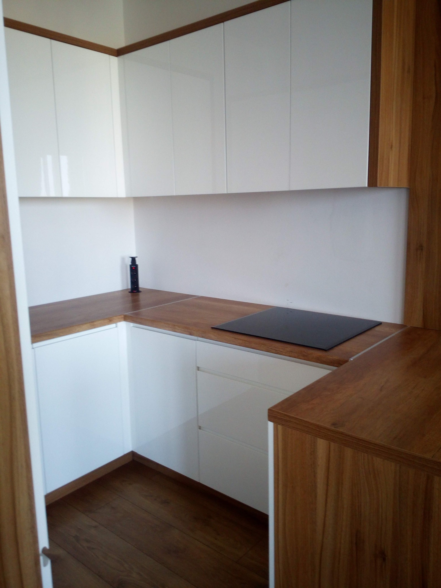 Nowoczesna, narożna zabudowa kuchenna z białymi, lakierowanymi szafkami i drewnianym blatem, płyta indukcyjna wkomponowana w blat, widoczny wysuwany kontakt blatowy.