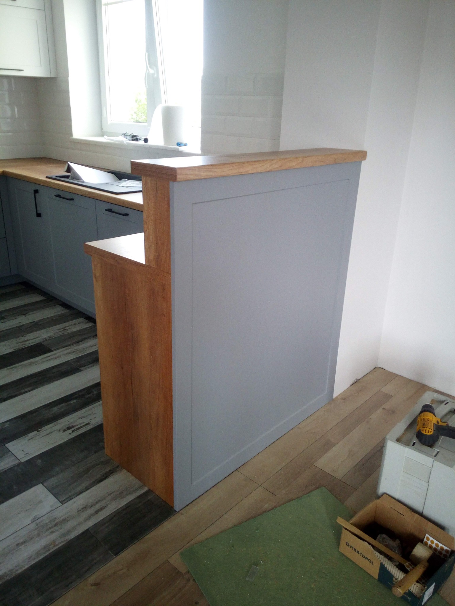 Szara zabudowa kuchenna z drewnianym blatem i elementami wykończeniowymi w trakcie montażu, widoczne narzędzia i materiały budowlane na podłodze.