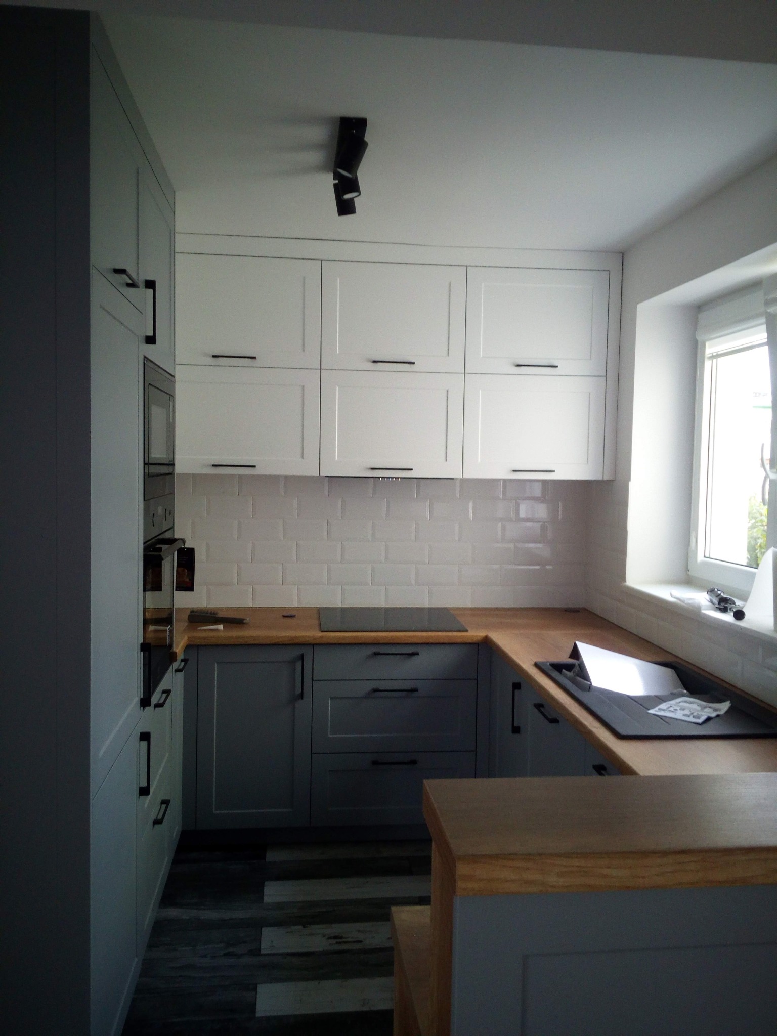 Nowoczesna kuchnia w odcieniach szarości i bieli z drewnianym blatem, płytki metro na ścianie, oświetlenie punktowe na suficie, widok na okno z roletą.