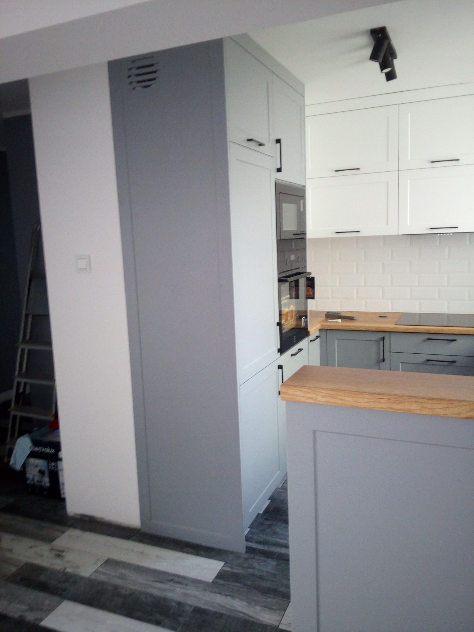Szara zabudowa kuchenna z piekarnikiem i mikrofalówką, białe szafki wiszące, drewniany blat, płytki na ścianie w stylu metro, widoczna drabina w tle, podłoga imitująca drewno.