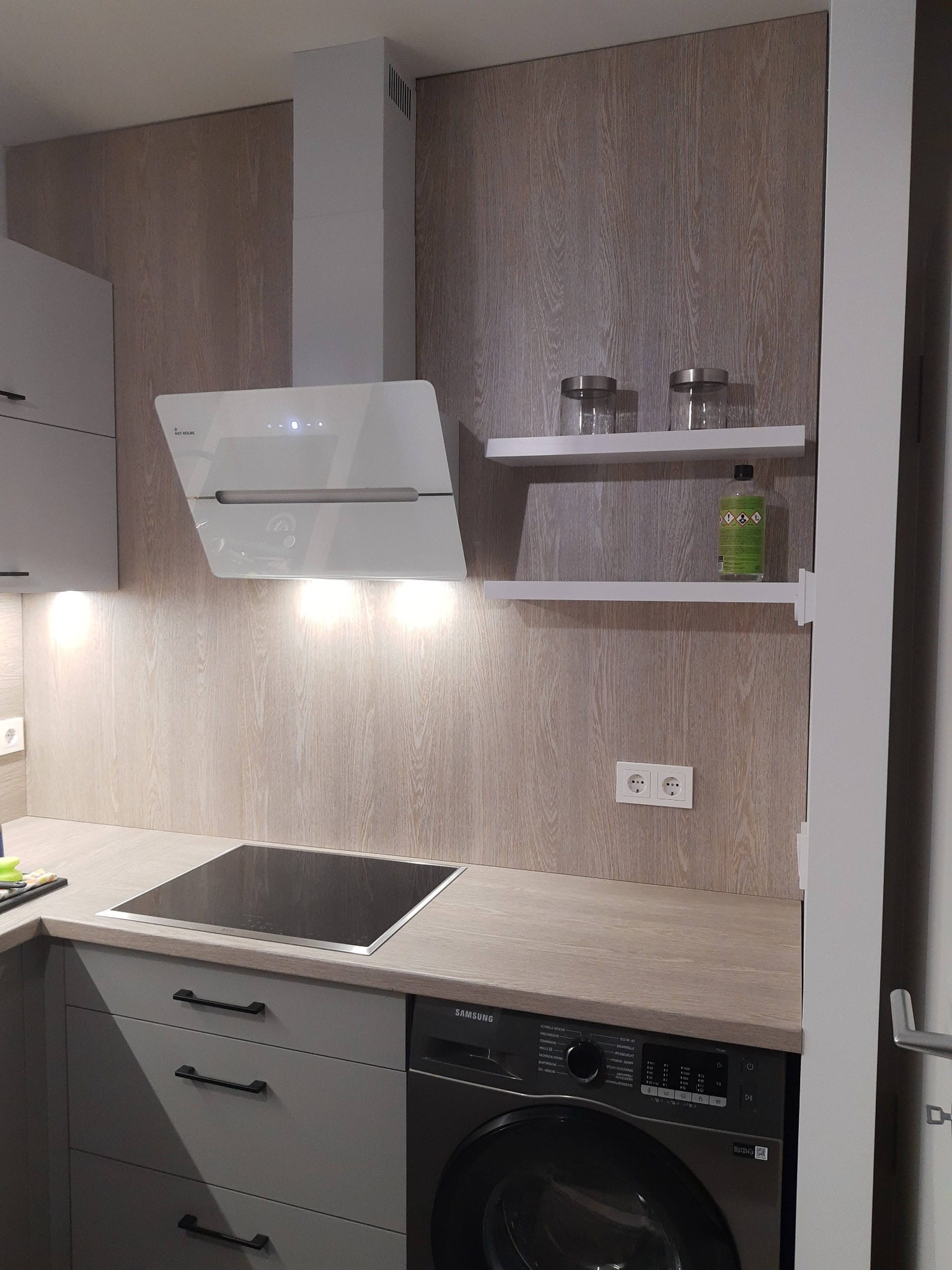 Szara kuchnia z drewnianym blatem i płytą indukcyjną, biały okap i półki z pojemnikami. Pralka zabudowana w szafce. Oświetlenie podszafkowe.