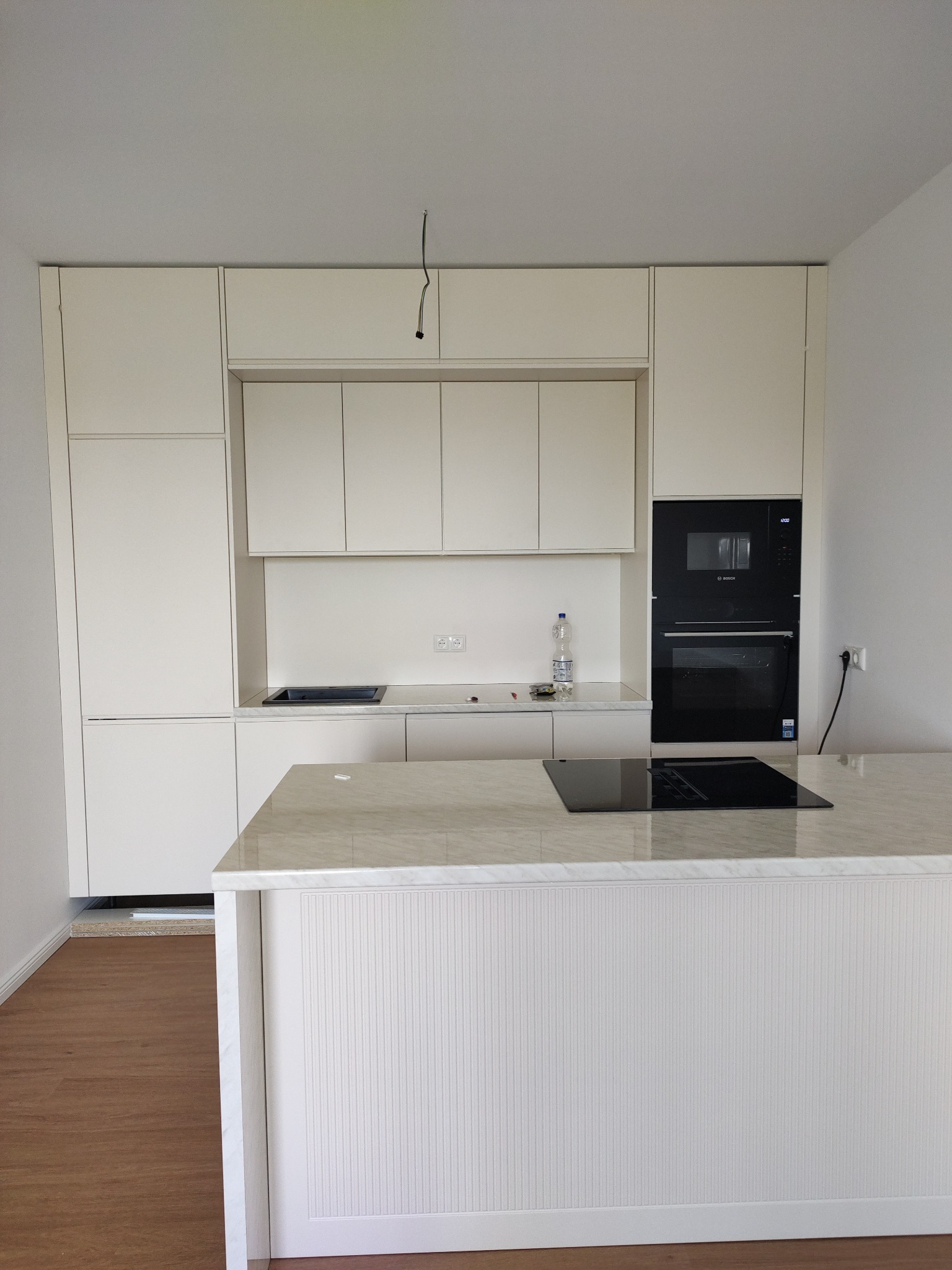 Jasna kuchnia z wyspą i marmurowym blatem. Widoczne wbudowane sprzęty AGD, płyta indukcyjna i minimalistyczne fronty szafek. Podłoga drewniana, oświetlenie sufitowe.