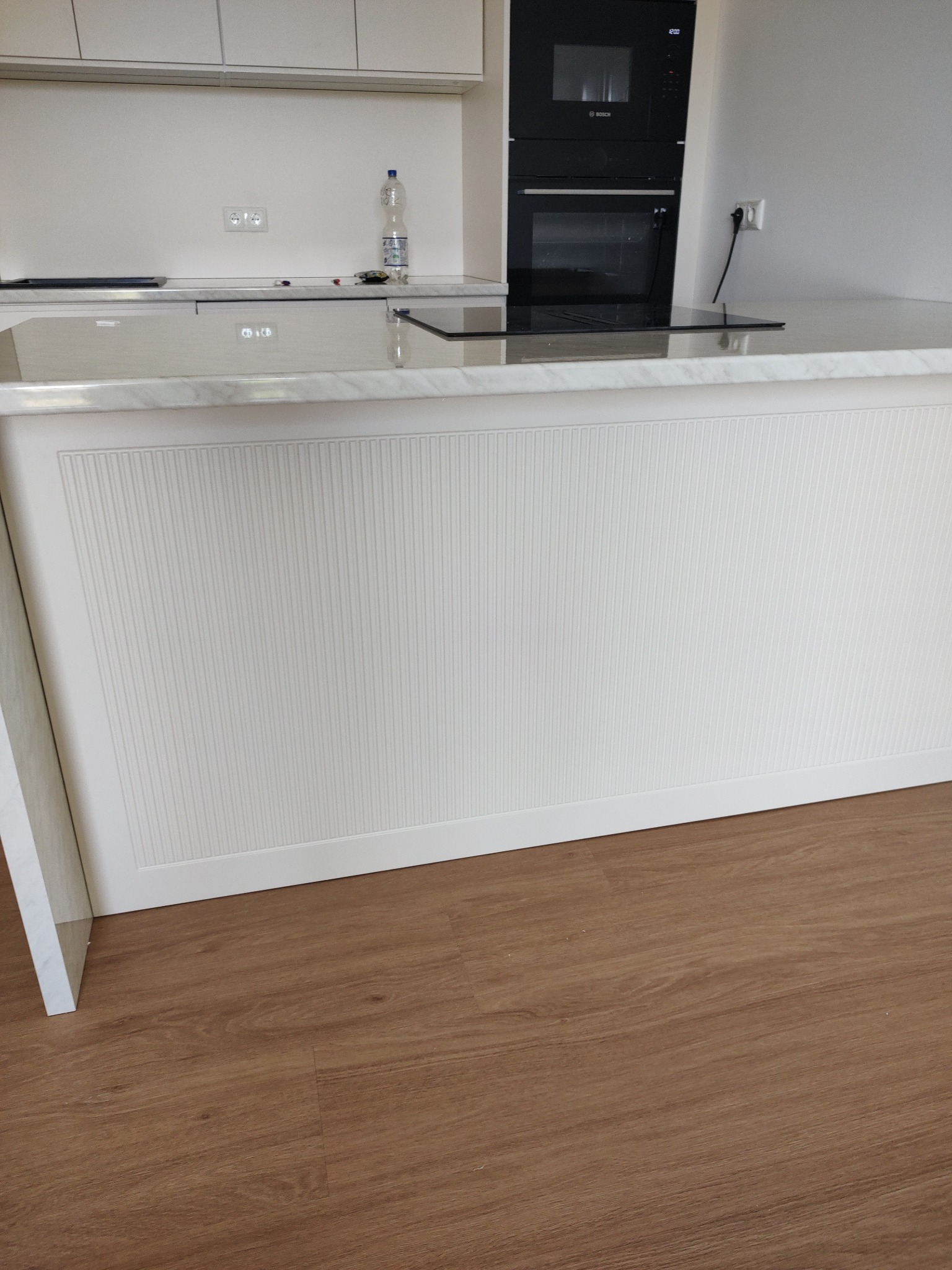 Biała wyspa kuchenna z frezowanymi frontami na tle zabudowy kuchennej z piekarnikiem Bosch i marmurowym blatem, na podłodze panele drewnopodobne.