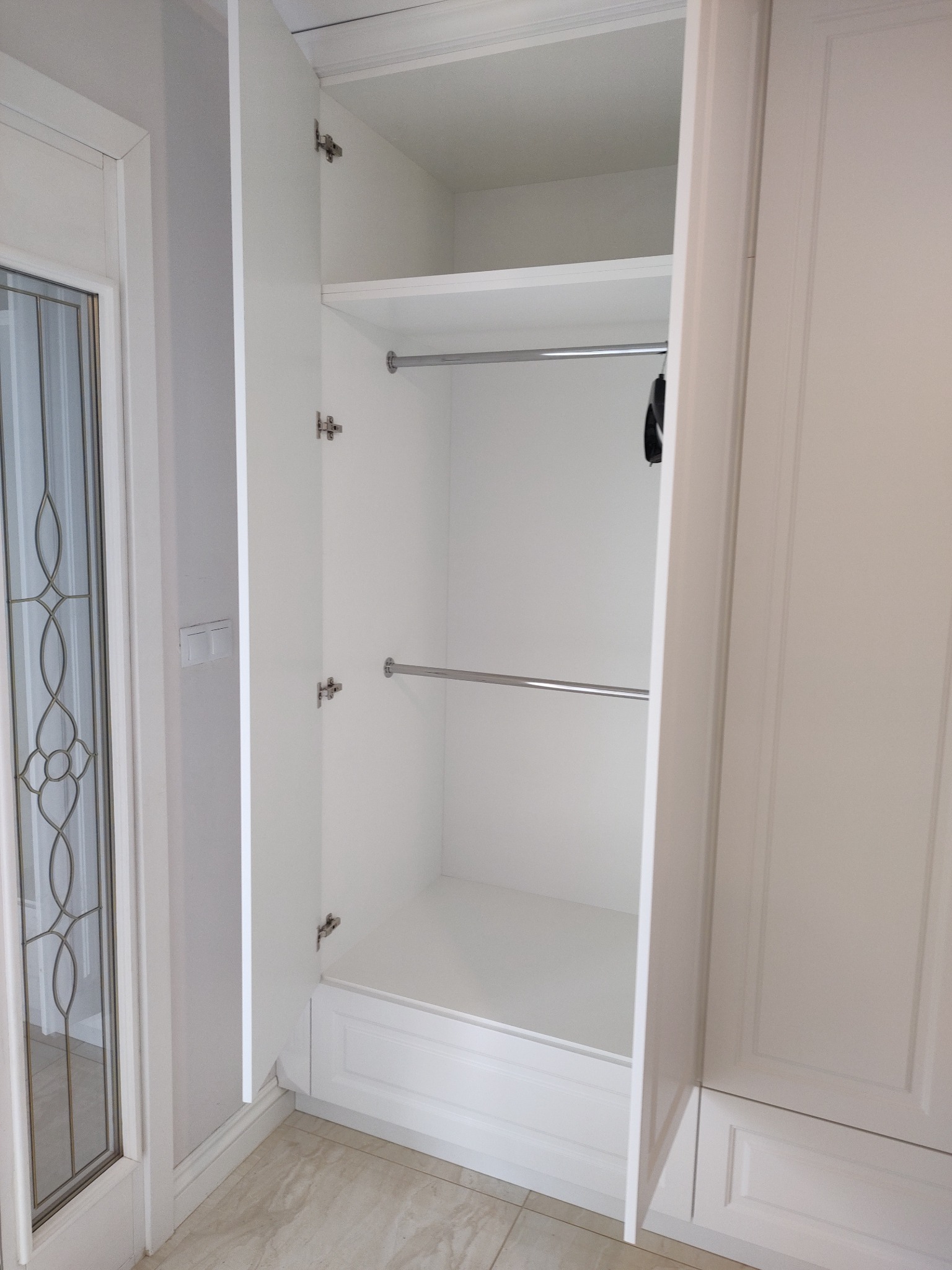 Otwarta, biała szafa wnękowa z półką i drążkami na ubrania, widoczna z perspektywy wnętrza. Minimalistyczny design, jasne wnętrze.