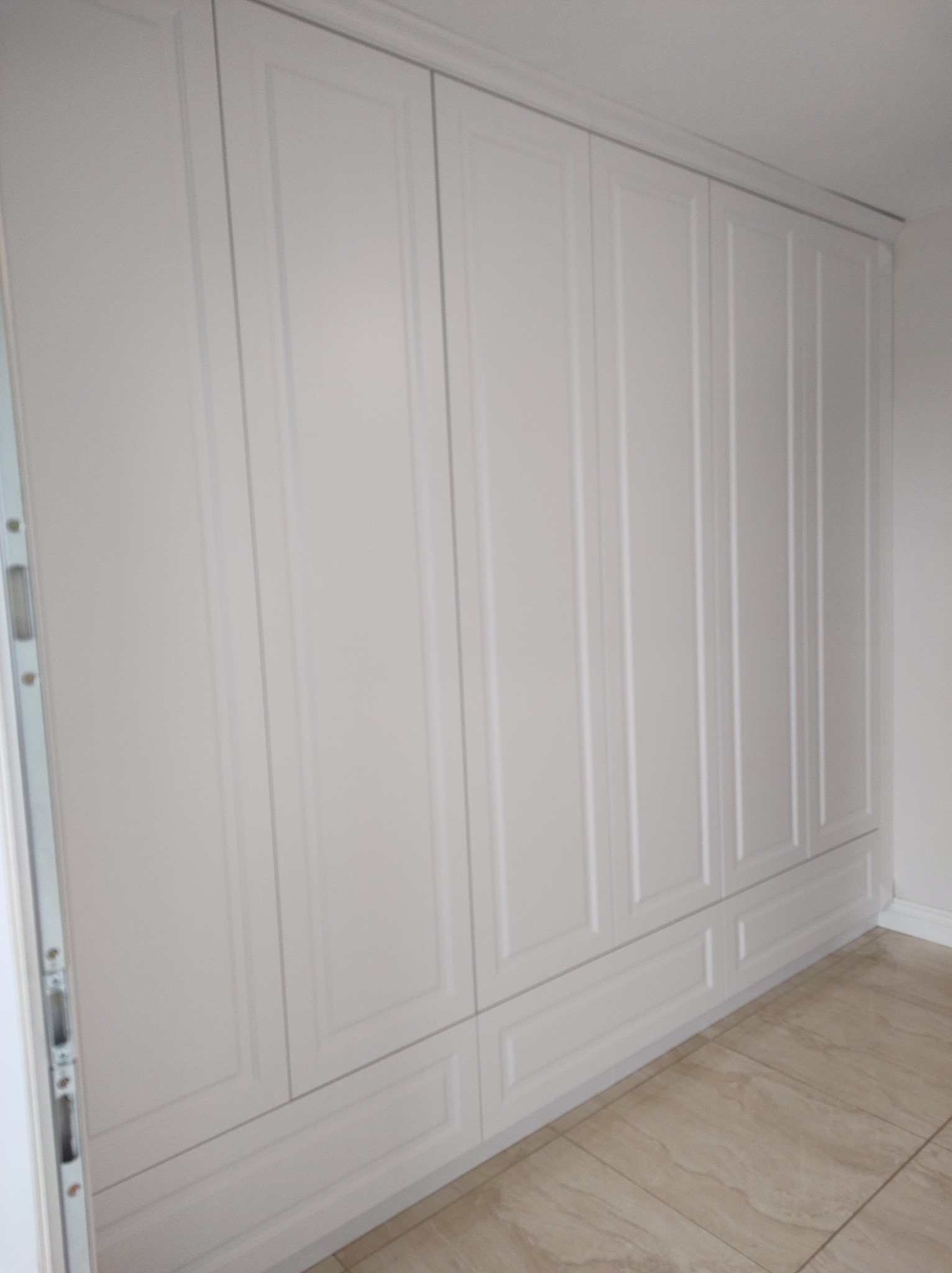 Biała, zabudowana szafa wnękowa z frezowanymi frontami, dopasowana do jasnego wnętrza. Widoczny fragment podłogi z płytek imitujących marmur.
