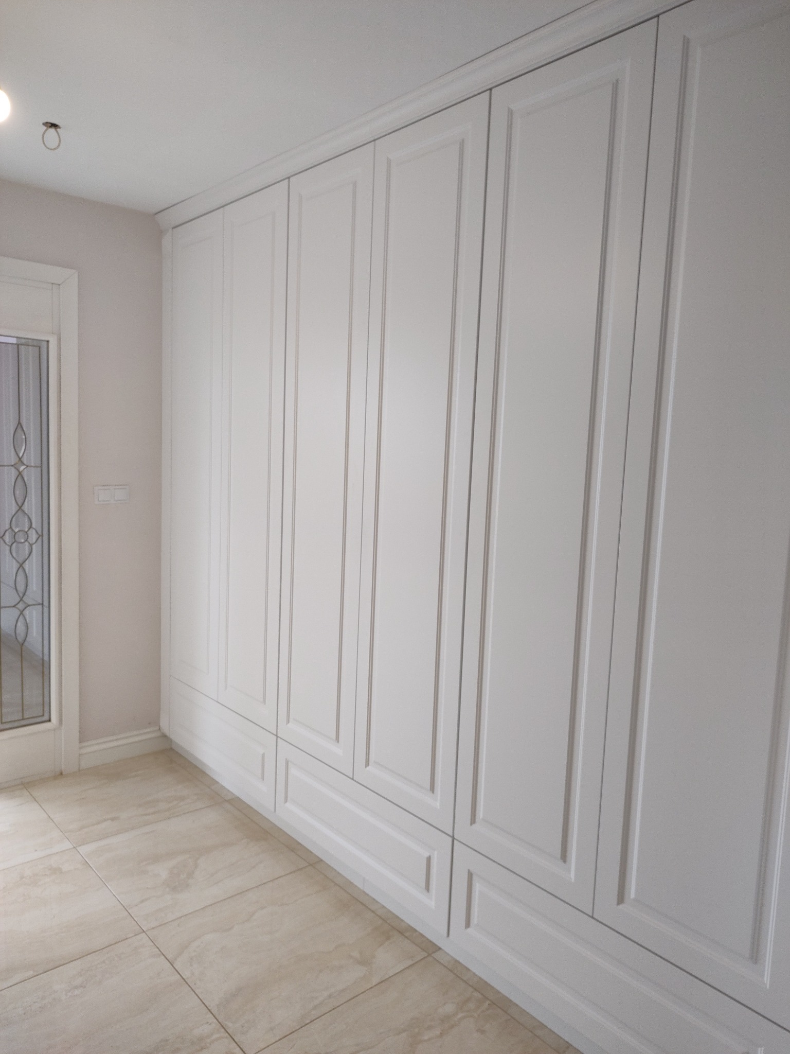 Biała szafa wnękowa z frezowanymi frontami, dopasowana do wysokości pomieszczenia, z dekoracyjną listwą przypodłogową i ozdobnymi drzwiami po lewej.
