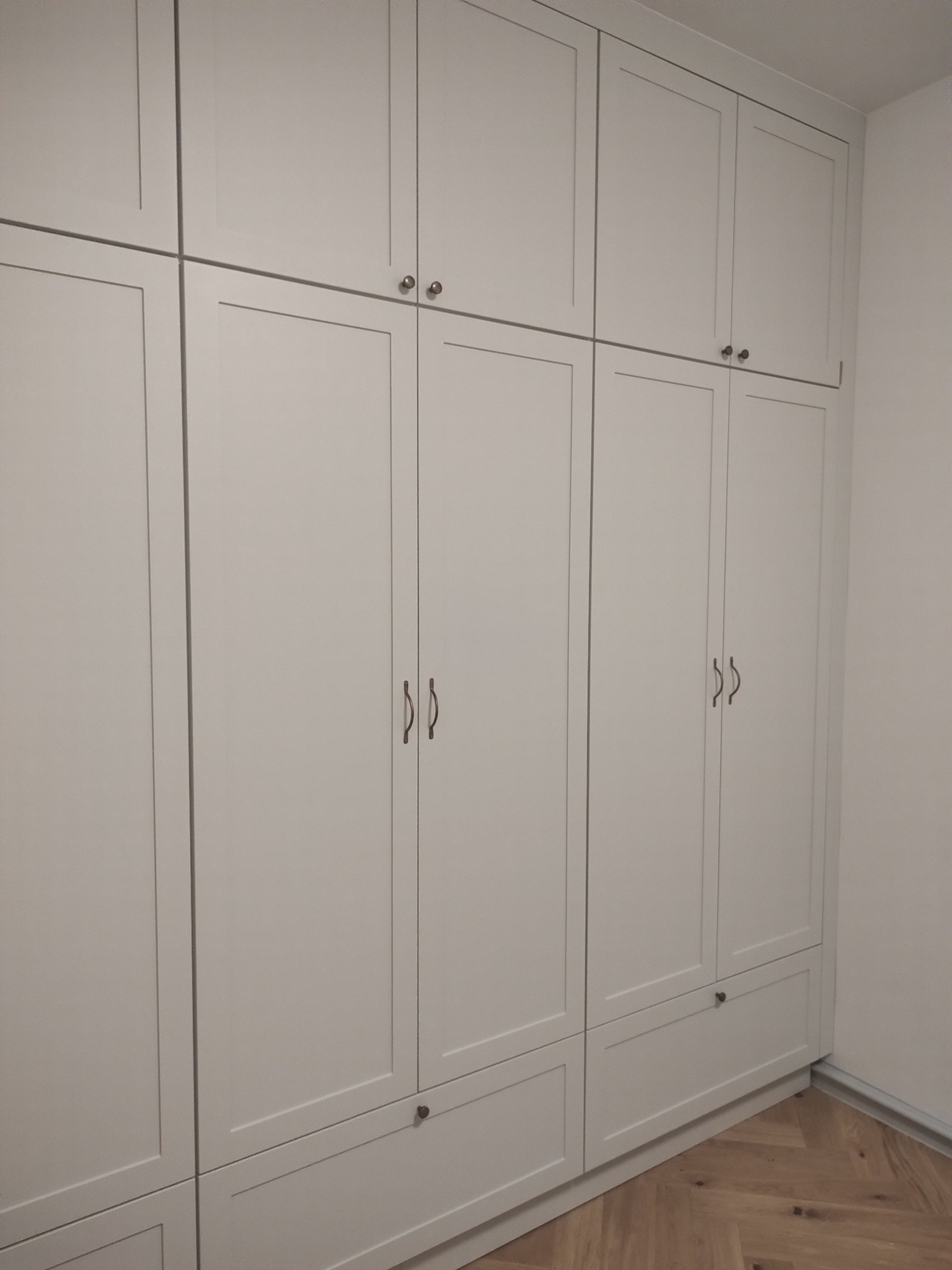 Biała, zabudowana szafa z szufladami i górnymi szafkami, z delikatnymi uchwytami, na tle jasnej ściany i drewnianej podłogi ułożonej w jodełkę.