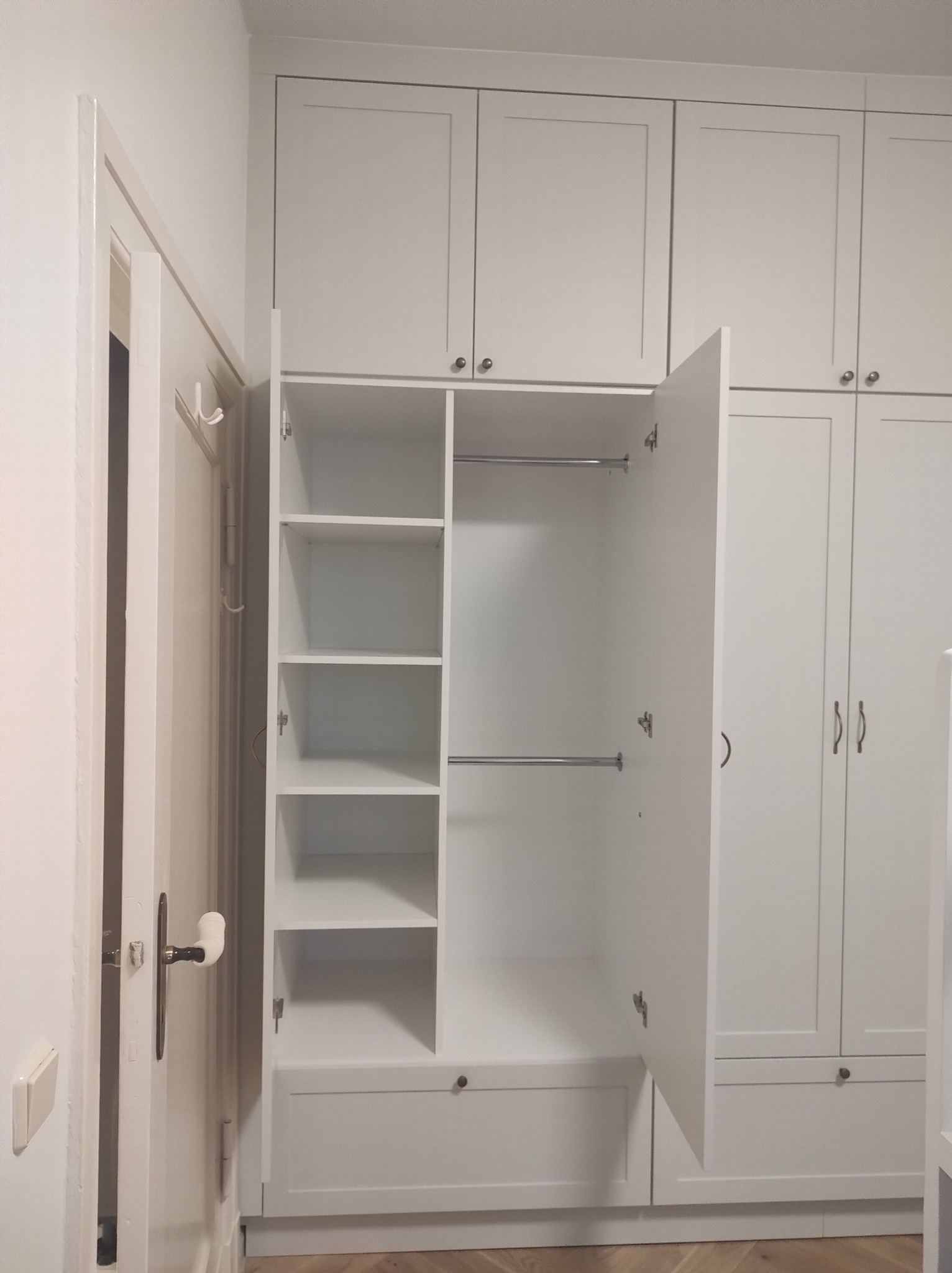 Biała szafa wnękowa z otwartymi drzwiami, widoczne półki i drążki na ubrania. Klasyczny design, jasne wnętrze. Ujęcie z bliska, skupione na wnętrzu szafy.