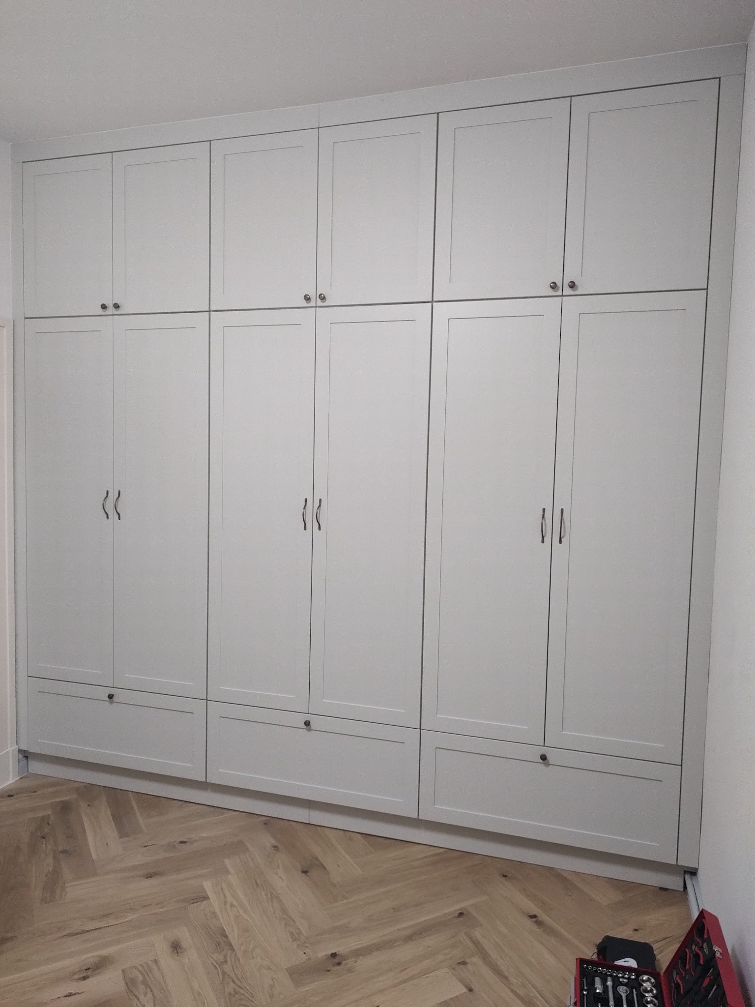 Jasnoszara, zabudowana szafa z górnymi szafkami i dolnymi szufladami, zamontowana na podłodze z parkietem w jodełkę. Widoczne narzędzia, sugerujące świeży montaż.