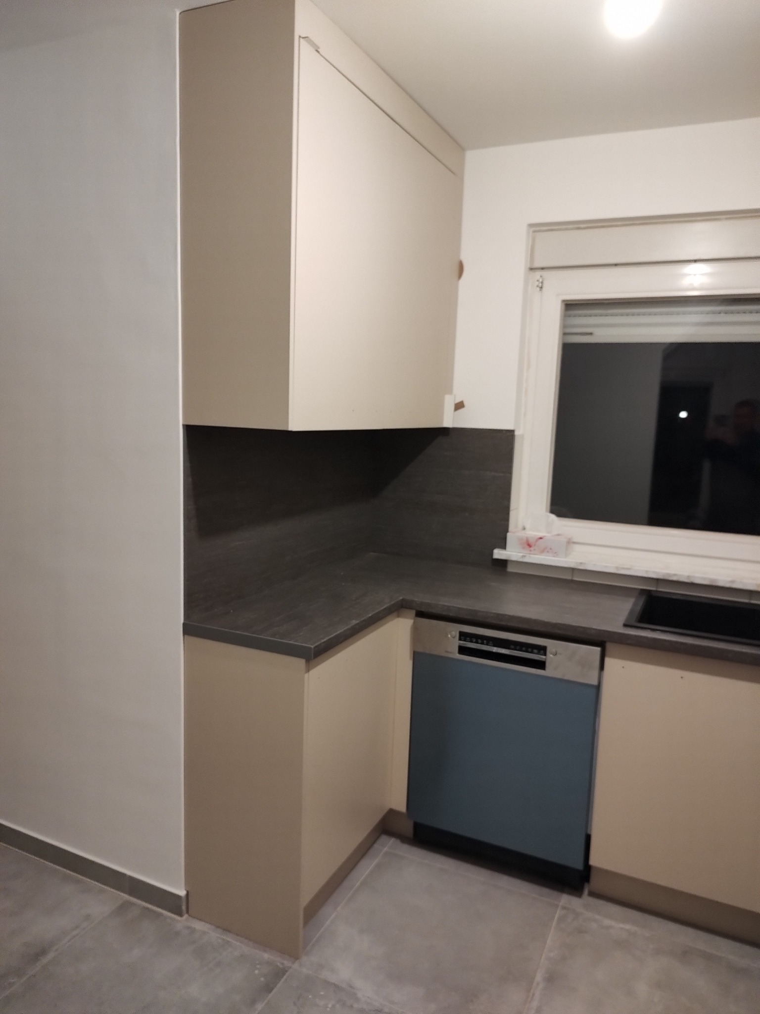 Jasna kuchnia z szarym blatem i niebieską zmywarką w zabudowie. Ujęcie z niskiego kąta, widoczny fragment okna i szare płytki podłogowe.