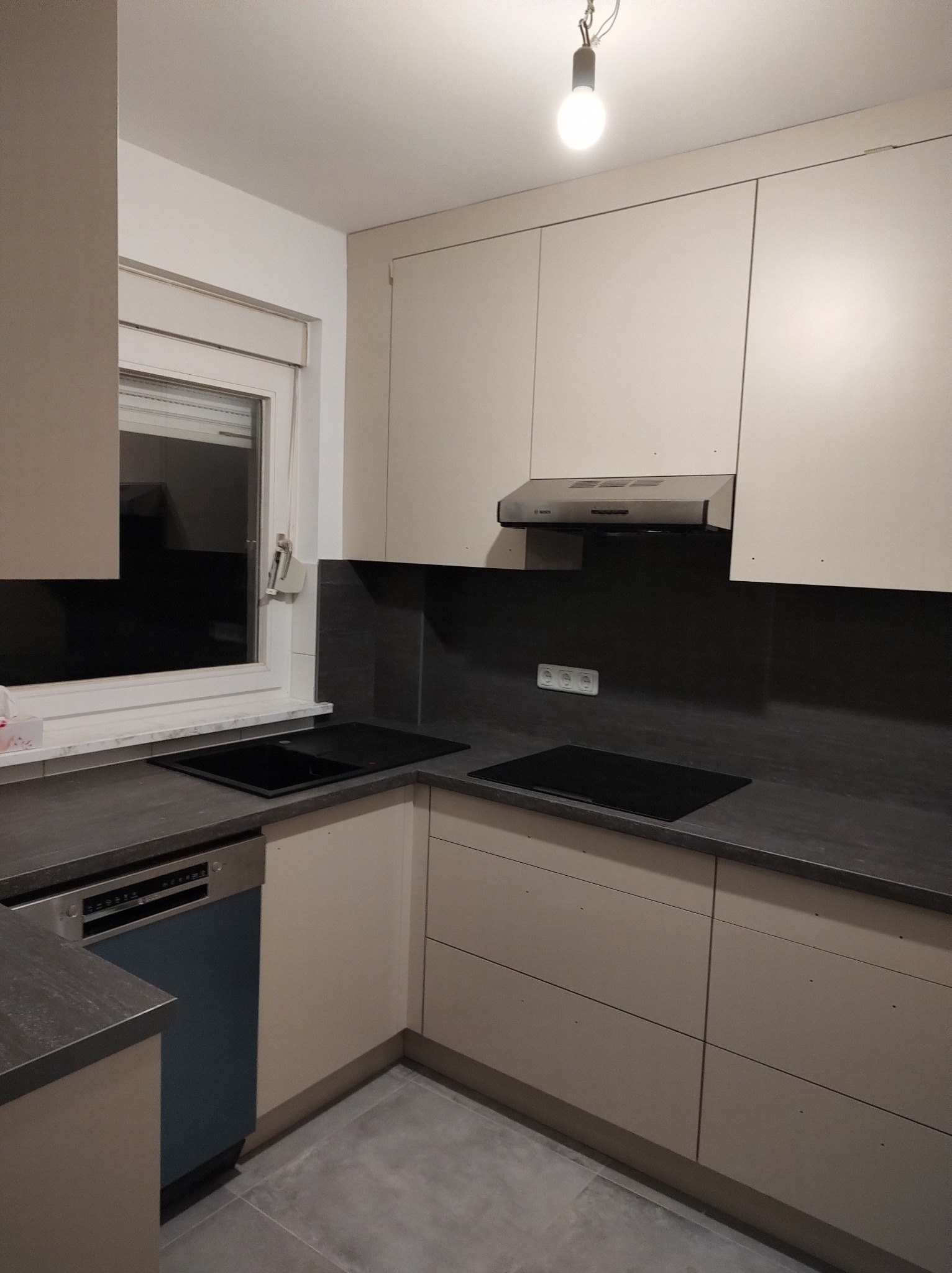 Jasna kuchnia z szarym blatem i niebieską zmywarką. Widoczna płyta indukcyjna, okap i gładkie fronty szafek. Minimalistyczny design, stonowana kolorystyka.