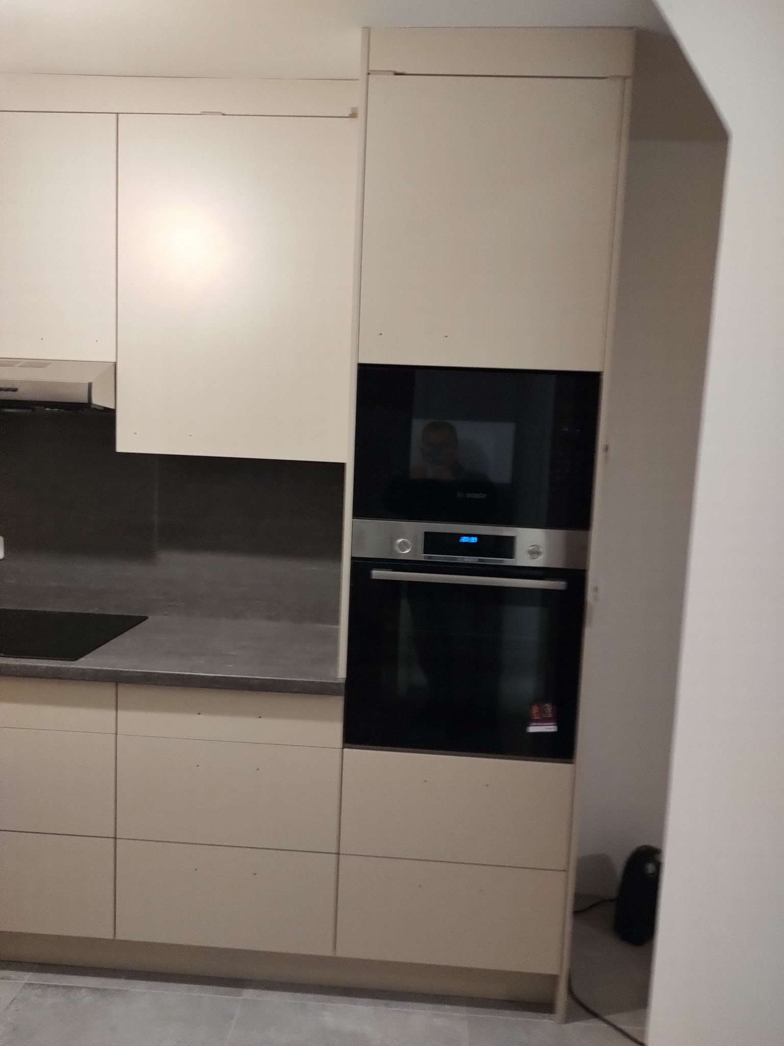 Jasna zabudowa kuchenna z piekarnikiem i płytą w Koninie. Minimalistyczne fronty szafek, szary blat i płytki nad blatem. Widoczna refleksja w szybie piekarnika.