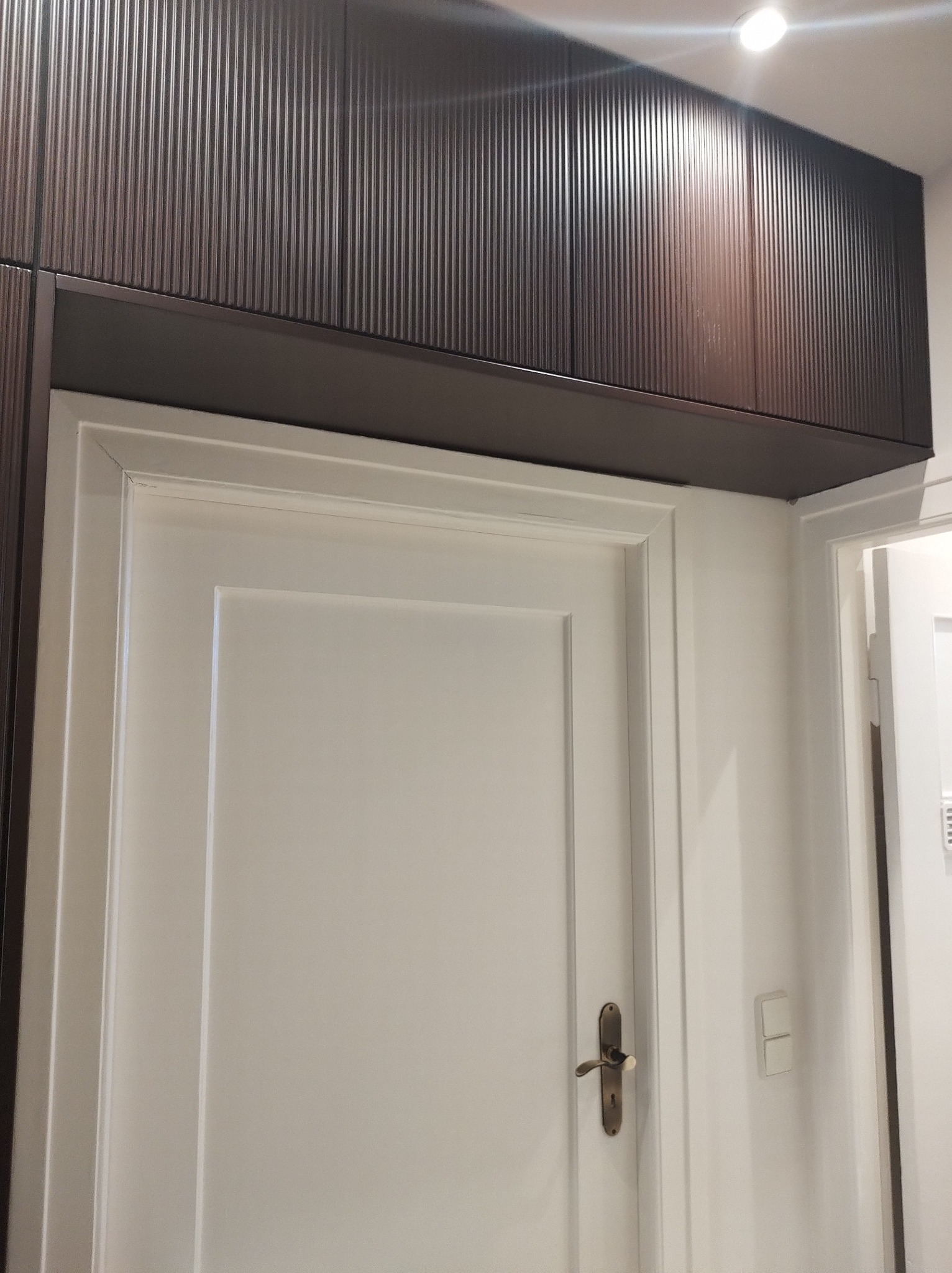 Białe drzwi wewnętrzne z brązową szafą wnękową nad futryną. Widoczne detale wykończenia i oświetlenie sufitowe.