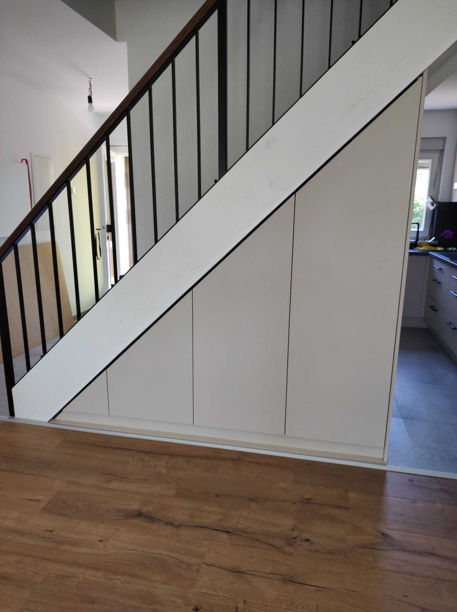 Szafa wnękowa pod schodami, z jasnymi frontami i subtelnymi uchwytami, idealnie wkomponowana w przestrzeń. Widoczny fragment podłogi z paneli.