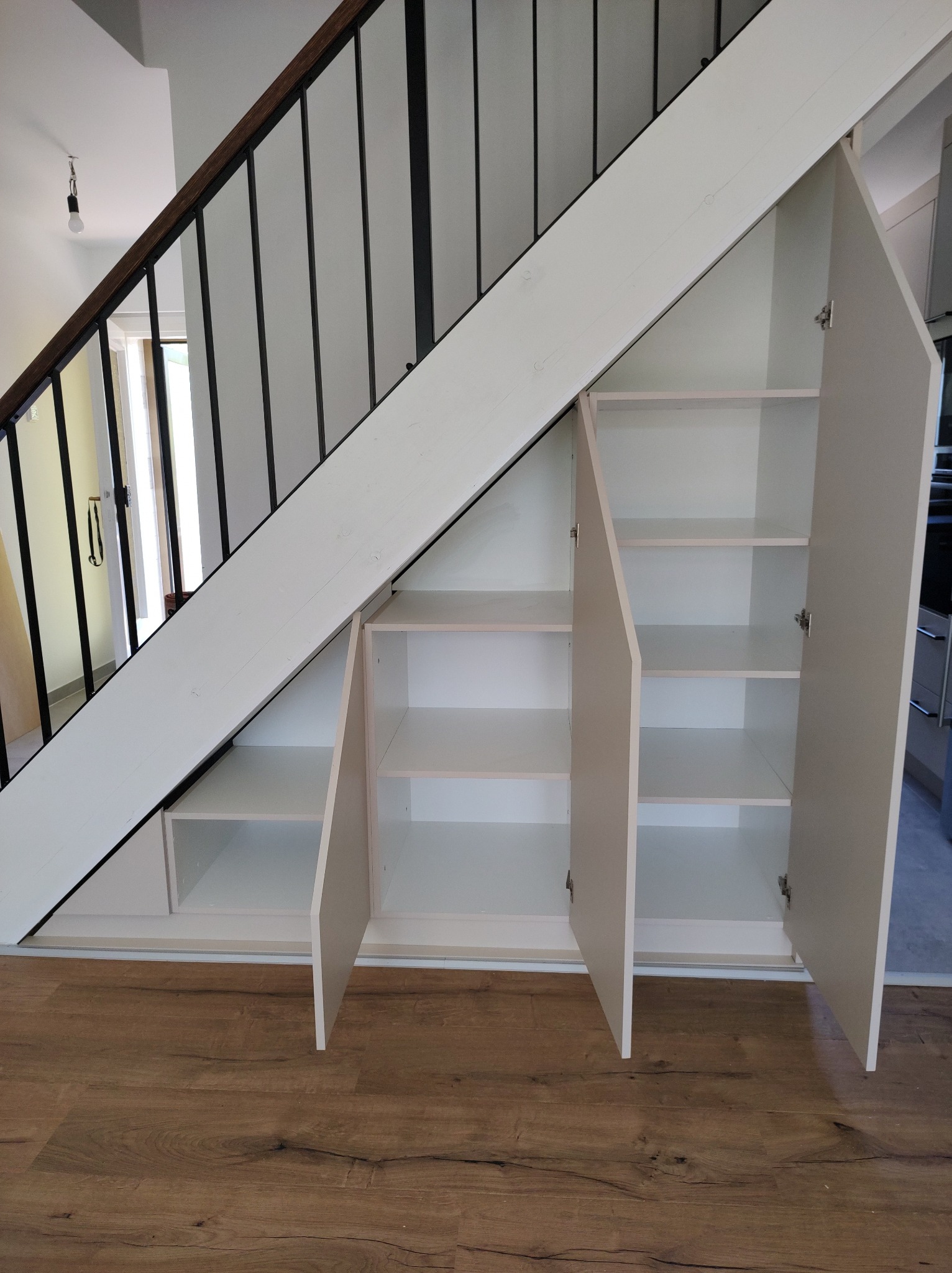 Biała szafa wnękowa pod schodami z otwartymi drzwiczkami ukazującymi półki, drewniana podłoga, czarne barierki schodowe.