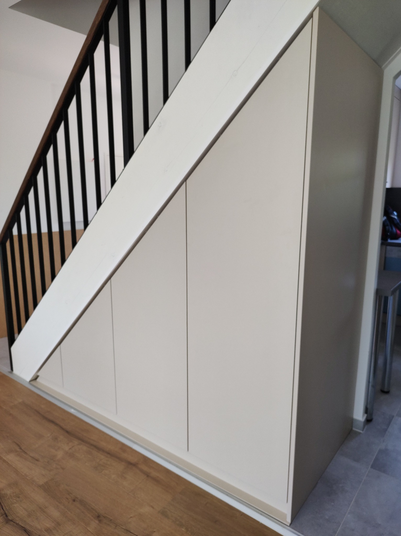 Biała szafa wnękowa pod schodami z czarną balustradą. Drewniana podłoga i szare płytki tworzą nowoczesny kontrast w jasnym wnętrzu.