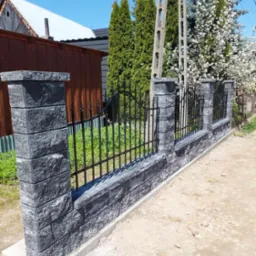 Nowoczesne ogrodzenie z szarej kostki łupanej z metalowymi przęsłami, widok na posesję z zielenią i kwitnącym drzewem, słup energetyczny w tle.