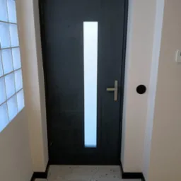 Nowoczesne drzwi wejściowe w kolorze grafitowym z pionowym przeszkleniem, zamontowane w jasnym wnętrzu z podłogą z lastryko i ścianą z luksferów.