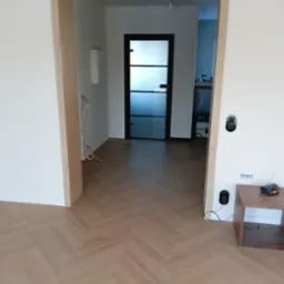 Ułożona podłoga z paneli w jodełkę francuską, widok na drzwi z czarną ramą i matowym szkłem, fragment ściany z gniazdkami i małym drewnianym stolikiem.