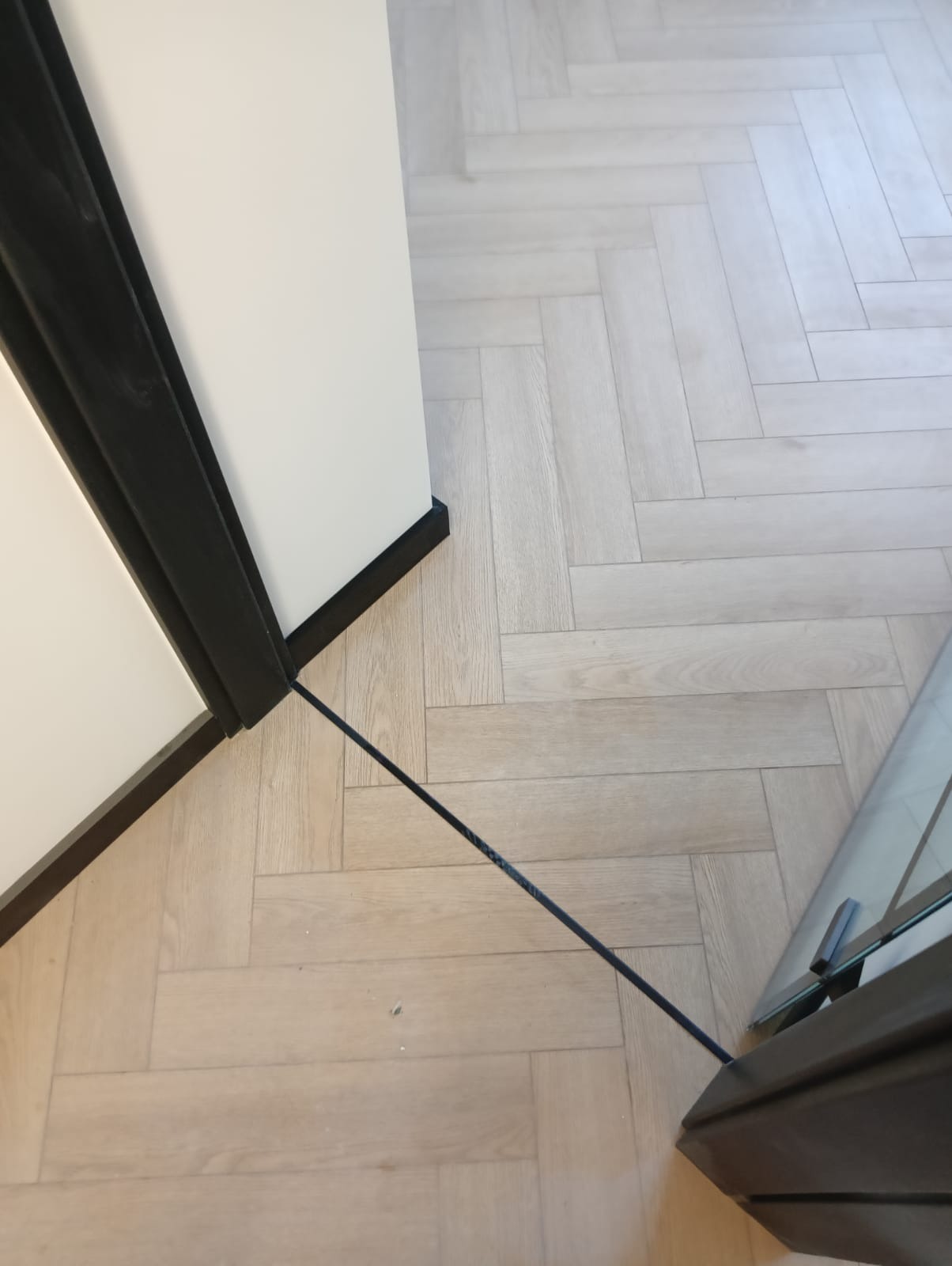 Jasna podłoga ułożona w jodełkę, z ciemnymi listwami przypodłogowymi i fragmentem kabiny prysznicowej z ciemną ramą.
