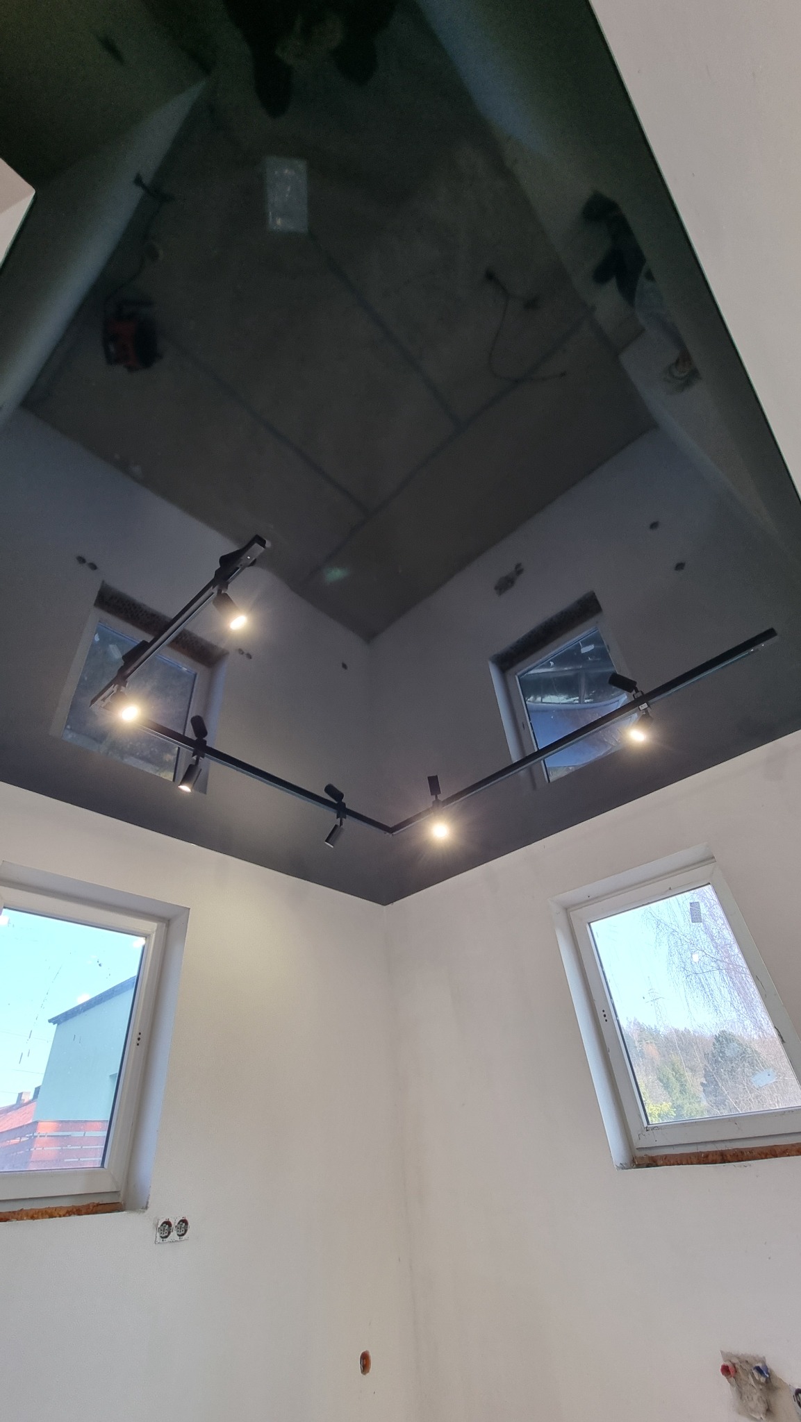 Czarny, błyszczący sufit napinany z zamontowanym systemem oświetlenia szynowego w narożnym pomieszczeniu z trzema oknami.