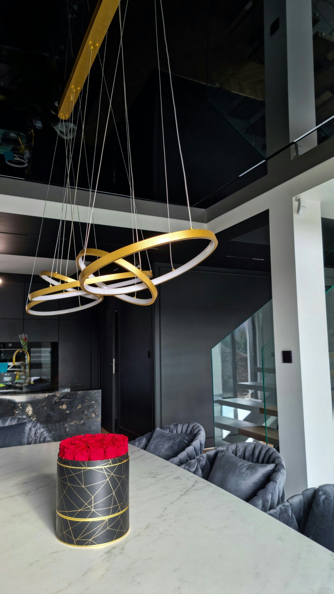 Nowoczesne wnętrze domu z czarnym sufitem napinanym, złotymi lampami pierścieniowymi, marmurowym stołem i szarymi krzesłami, dekorowane pudełkiem czerwonych róż.