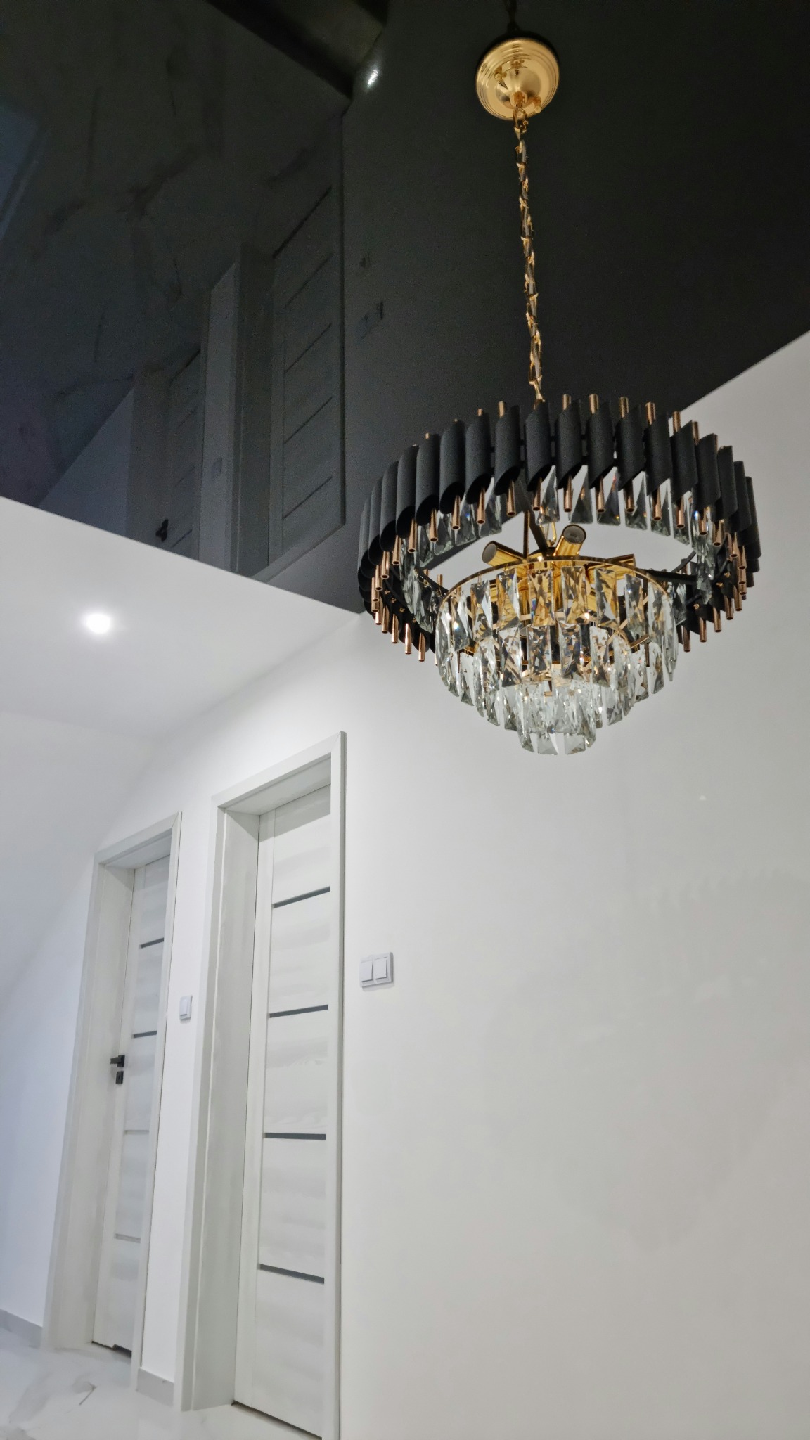 Czarny, błyszczący sufit napinany z nowoczesnym, okrągłym żyrandolem z kryształkami i złotymi elementami, białe ściany i drzwi w tle.