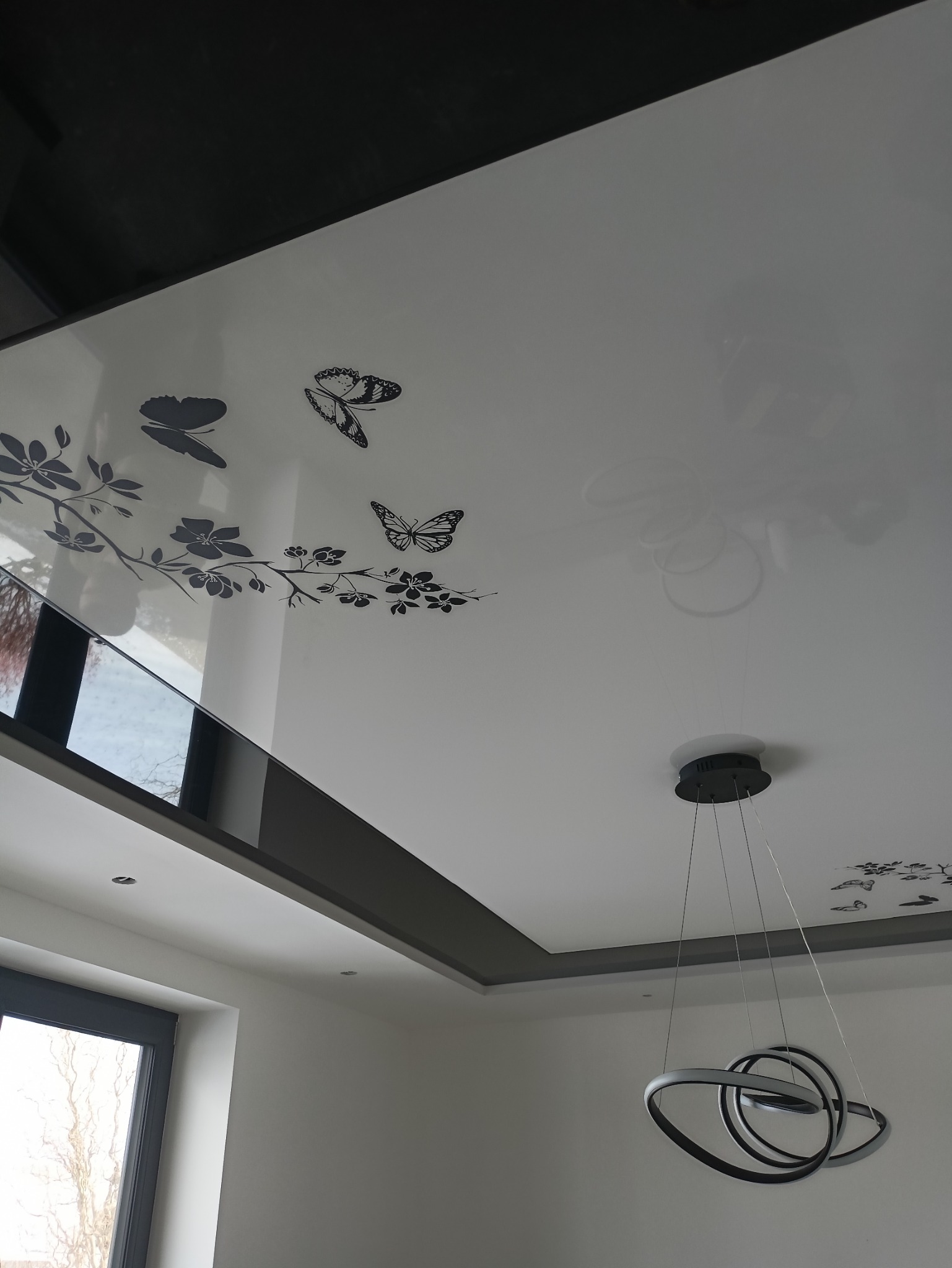 Biały, błyszczący sufit napinany z motywem czarnych motyli i kwiatów, nowoczesna lampa wisząca z okrągłymi elementami, szara rama sufitowa.