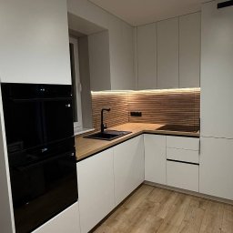 Aliaksandr Bortnik - Nowoczesna kuchnia z białymi szafkami, drewnianym blatem i panelem ściennym. Czarna armatura i sprzęt AGD. Oświetlenie LED podkreśla ciepły charakter wnętrza.