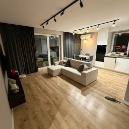 Aliaksandr Bortnik - Jasne panele podłogowe w salonie z beżową sofą narożną, czarnymi zasłonami i oświetleniem szynowym. Widok na aneks kuchenny z białymi szafkami.