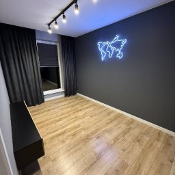 Aliaksandr Bortnik - Pokój po remoncie z drewnianą podłogą, szarą ścianą z neonową mapą świata i czarnymi zasłonami. Oświetlenie sufitowe punktowe. Minimalistyczny styl.
