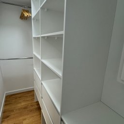 Aliaksandr Bortnik - Biała, zabudowana garderoba z półkami i szufladami, widok z dołu, drewniana podłoga, w tle wieszaki na ubrania. Nowoczesne wykończenie wnętrza.