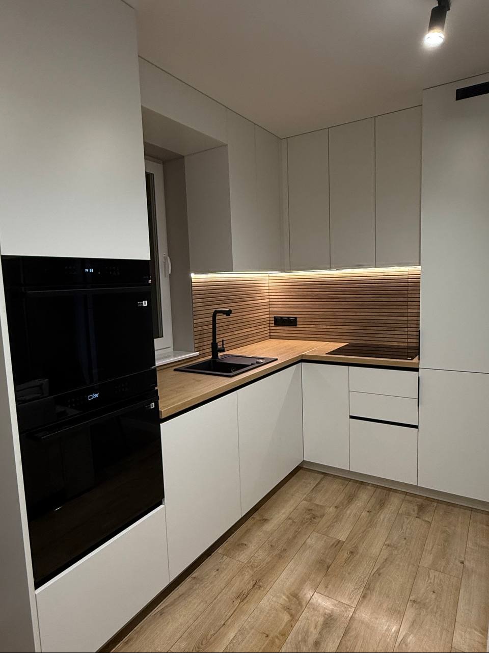 Nowoczesna kuchnia z białymi szafkami, drewnianym blatem i panelem ściennym. Czarna armatura i sprzęt AGD. Oświetlenie LED podkreśla ciepły charakter wnętrza.