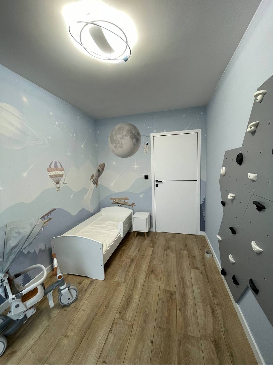 Pokój dziecięcy z motywem kosmicznym: fototapeta z planetami, łóżko, ścianka wspinaczkowa i drewniana podłoga. Nowoczesne oświetlenie sufitowe.