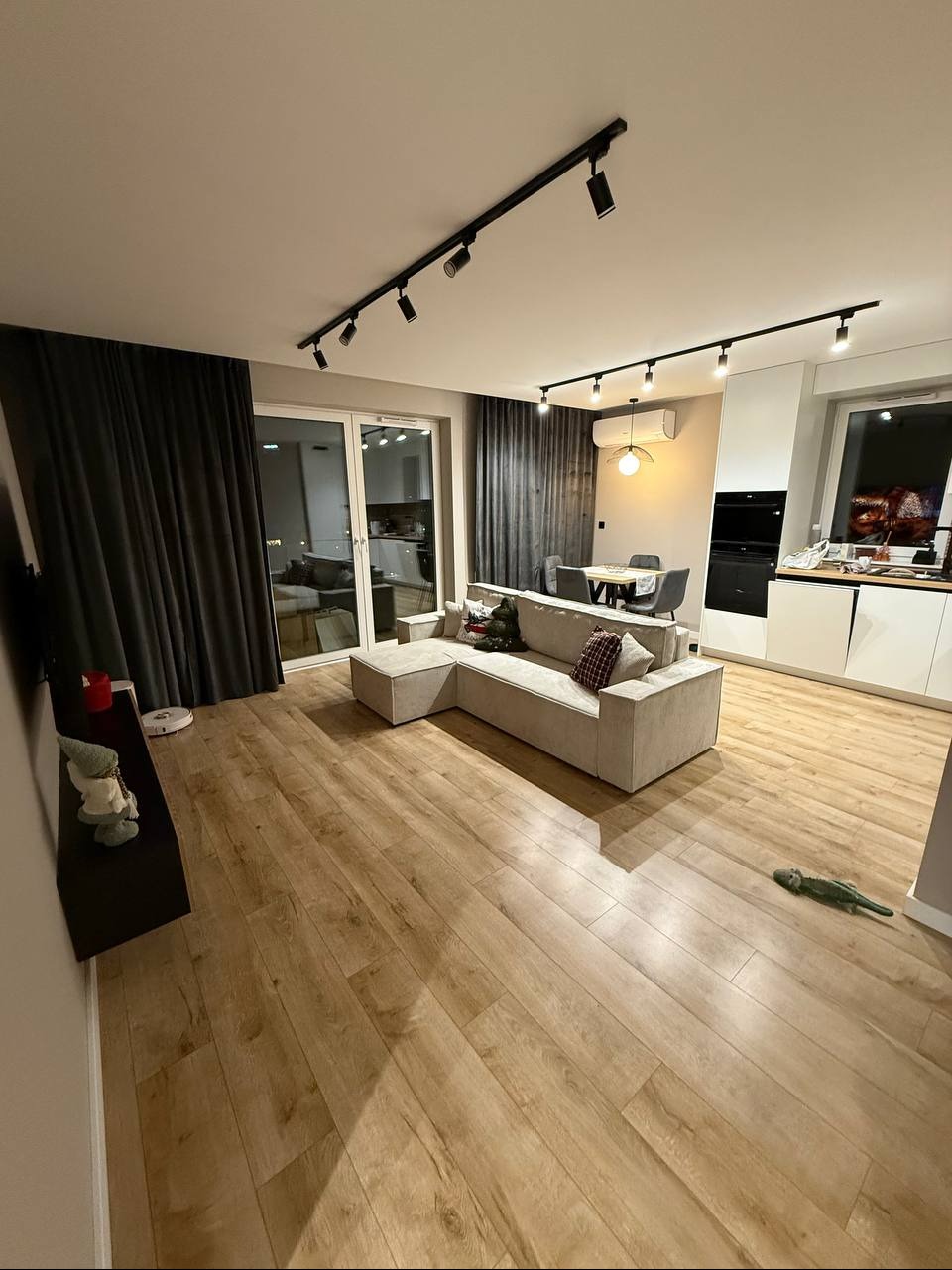 Jasne panele podłogowe w salonie z beżową sofą narożną, czarnymi zasłonami i oświetleniem szynowym. Widok na aneks kuchenny z białymi szafkami.
