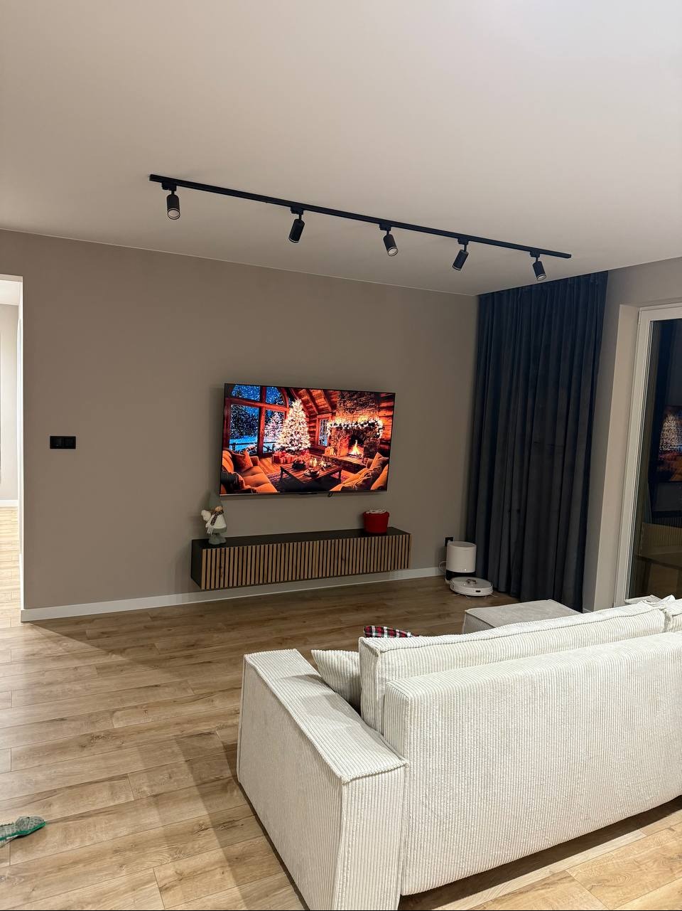 Nowoczesny salon z drewnianą podłogą, szarą ścianą i telewizorem z motywem świątecznym. Oświetlenie szynowe i minimalistyczna szafka RTV dodają elegancji.