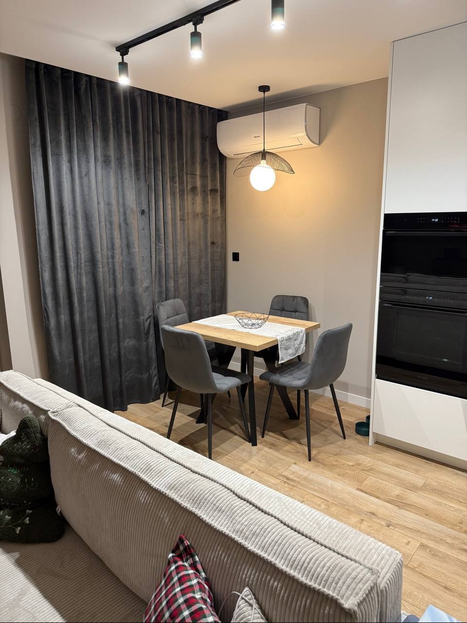 Nowoczesna jadalnia: drewniany stół z szarymi krzesłami, minimalistyczna lampa wisząca, szare zasłony i fragment sofy w tle. Klimatyczne oświetlenie.