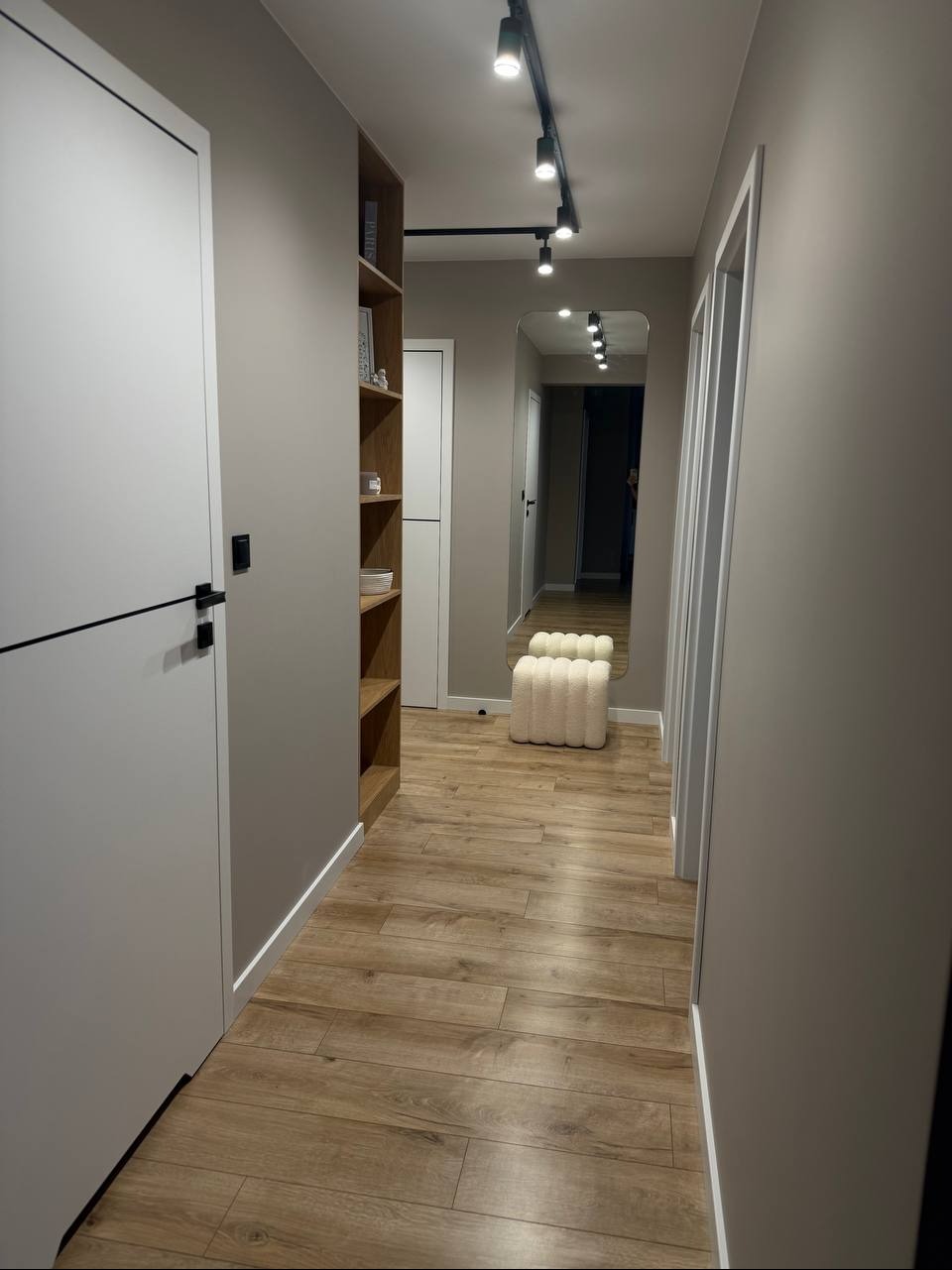 Nowoczesny przedpokój z drewnianą podłogą, białymi drzwiami z czarnymi akcentami, półkami i lustrem. Oświetlenie punktowe na suficie. Minimalistyczny design.